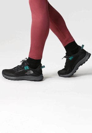 Zapatillas deportivas negras con un upper texturizado, que presentan acentos en color azul verdoso, una suela resistente y un logo en el lateral. Utilizadas con leggings rojos.