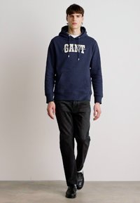 Felpa con cappuccio blu navy con logo "GANT", abbinata a jeans scuri e scarpe nere. Presenta lacci e tasca frontale. Texture liscia.