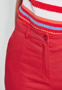 Pantalon en coton rouge avec une texture lisse, doté d'une fermeture à bouton et de poches latérales. Associé à un haut en maille rayé multicolore.