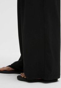 Pantalons larges noirs en tissu lisse, associés à des tongs noires. Les pantalons présentent une coupe droite et un ourlet net.
