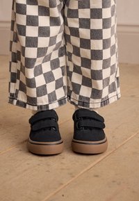 Melni velcro apavi ar teksturētu auduma virsu un brūnām gumijas zolītēm, kopā ar melnas un baltas krāsas rūtainām biksēm. Fona plakans koka grīdas segums.