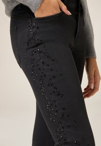 Jeans negros con un diseño ajustado, adornados con lentejuelas negras dispuestas en un patrón vertical a lo largo del muslo exterior.