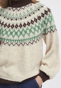 Knit sweater in cream with a geometric yoke pattern in green and brown. Features a high neck and ribbed brown cuffs and hem.
