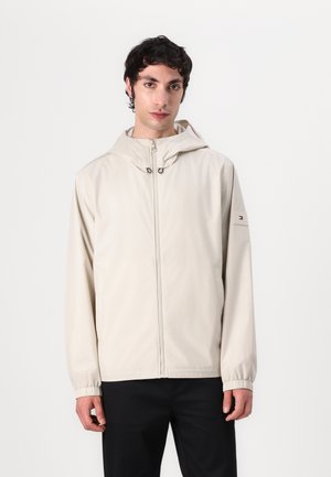 Lys beige hættejakke lavet af glat stof. Har en frontlynlås, elastiske manchetter og et logo patch på venstre ærme.