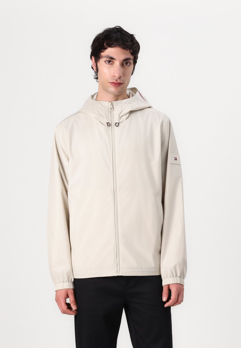 Light beige hooded jacket made from smooth fabric. Features a front zipper, elastic cuffs, and a logo patch on the left sleeve.