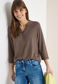 Femme souriante, portant une chemise marron à boutons aux manches trois-quarts et un jean bleu, tenant un tissu jaune dans sa main droite.