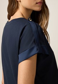 Blusa azul marino de mangas cortas, con un pliegue de satén en contraste y un botón decorativo dorado en el hombro. Textura suave.