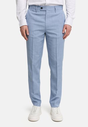 Man draagt lichtblauwe slim-fit nette broek met een wit overhemd en witte sneakers, staat met ontspannen handen langs zijn zij.