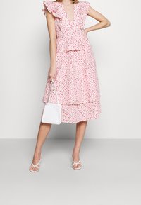 Vaaleanpunainen kukkakuvioinen mekko, jossa on röyhelöhihat, porrastettu malli, kevyt kangas ja syvä pääntie; yhdistetty pieneen valkoiseen käsilaukkuun ja sandaaleihin.