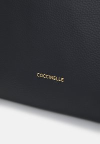 Sac en cuir bleu marine présentant une surface texturée avec un détail logo "Coccinelle" doré près du bas. Design minimaliste et finition élégante.