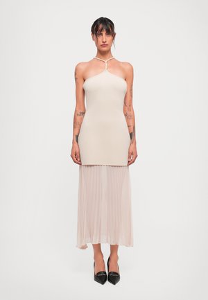 Femme avec des tatouages portant un top dos nu beige et une jupe plissée, se tenant debout avec les bras détendus, chaussée de talons aiguille noirs.