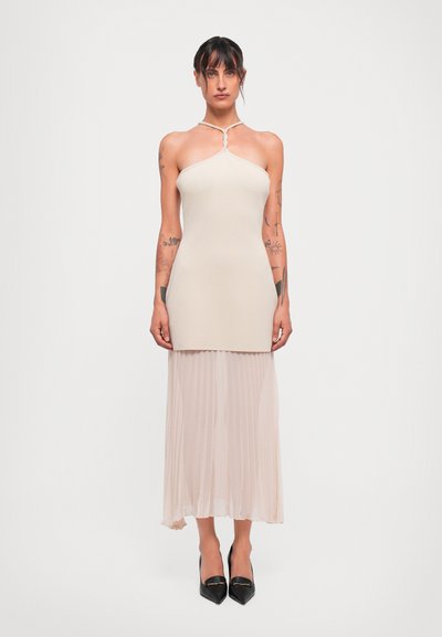 Femme avec des tatouages portant un top dos nu beige et une jupe plissée, se tenant debout avec les bras détendus, chaussée de talons aiguille noirs.