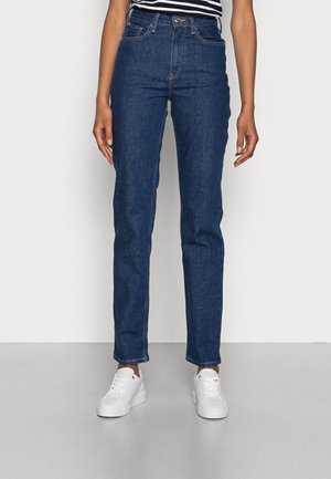 Personne portant un jean bleu foncé taille haute à jambes droites et des baskets blanches, debout devant un fond uni clair.