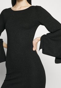 Robe noire en stretch avec de longues manches évasées. Tissu texturé avec une subtile brillance. Col rond et silhouette ajustée.