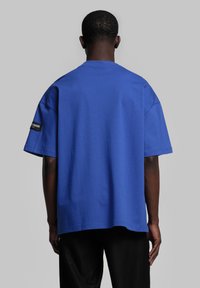 T-shirt en coton bleu surdimensionné avec un col rond, des manches courtes et un détail d'étiquette noire sur le bras gauche. Texture lisse, coupe décontractée.