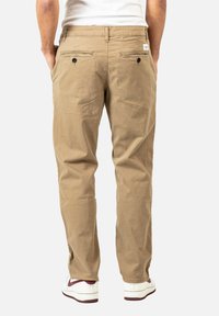Beige Baumwollhose mit lockerem Schnitt, ausgestattet mit zwei Gesäßtaschen und Knopfverschlüssen. Getragen mit weißen und burgunderfarbenen Sneakers.