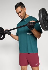 Téglakék Puma környakú póló fehér logóval, bordó rövidnadrággal, amelyen fehér szöveg látható. A felhasználó egy rúddal a kezében, amelyen fekete súlyok vannak.