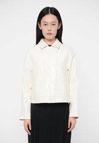 Γυναίκα με μακριά μαύρα μαλλιά, φορώντας ένα κρεμ μπουφάν με κουμπιά και γιακά, και μαύρο παντελόνι, που στέκεται μπροστά σε λευκό φόντο.