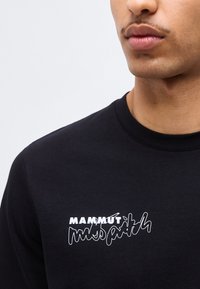 Κοντινή λήψη ενός ατόμου που φοράει μαύρο πουκάμισο με λευκή επιγραφή "Mammut mispath" στο στήθος, εμφανίζοντας τα χείλη και το γραμμή της γνάθου.