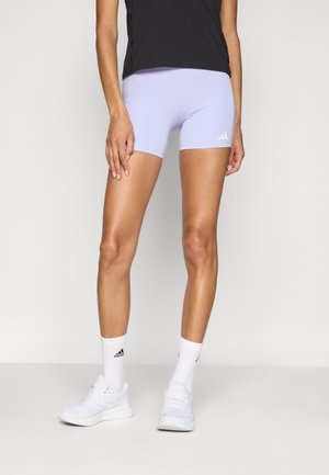 Žena nosící světle fialové sportovní šortky, černý top, bílé ponožky Adidas a bílé tenisky stojí na jednobarevném pozadí.