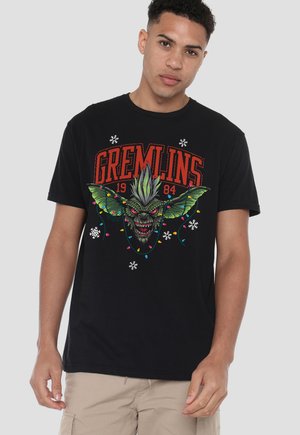 Young man wearing black t-shirt with colorful Gremlins character, Christmas lights, snowflakes, and "GREMLINS 1984" text in red.