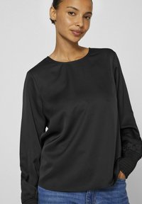 Blusa nera a maniche lunghe in tessuto liscio, con collo tondo e polsini arricciati. Abbinata a jeans in denim blu.