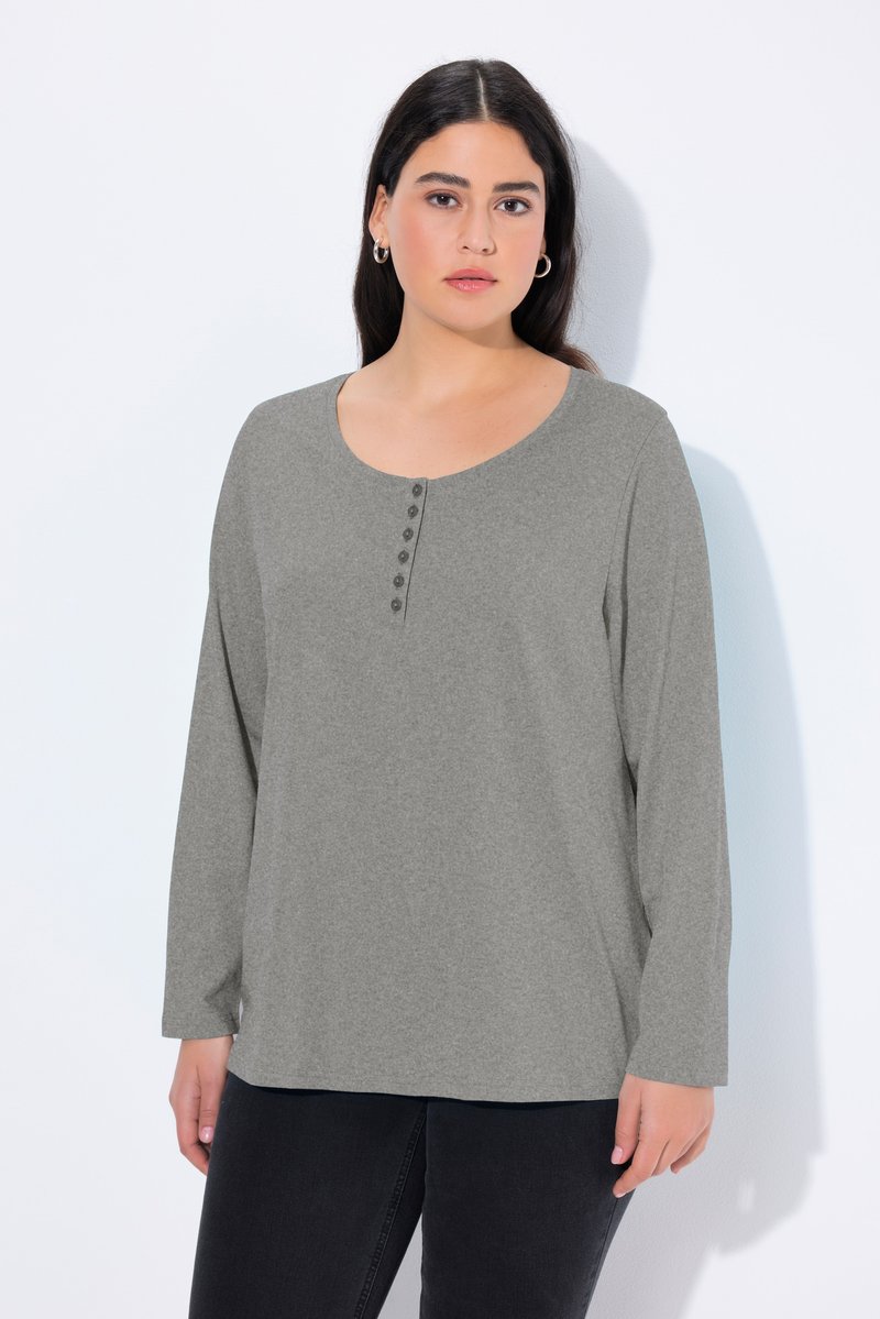 Mujer con pelo largo y oscuro, vestida con un top gris de manga larga con botones delanteros y pantalones negros, de pie frente a un fondo blanco.
