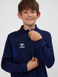 Jongen lacht terwijl hij een marineblauwe Hummel sportjas dichtritst tegen een effen lichte achtergrond.