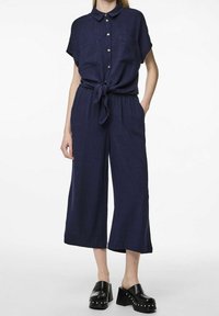 Tuta blu navy a maniche corte con chiusura a bottoni, vita con cintura e due tasche frontali. Il tessuto sembra leggero e testurizzato, con gambe larghe.