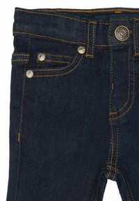 Mörkblå denimjeans med guldstickning, femfickorsdesign och metalldetaljer för förslutning. Närbild av midjebandet och framfickan.