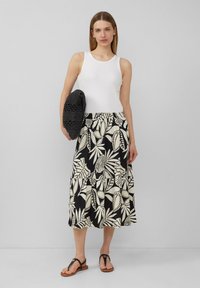 Schwarz-weißer, gestufter Rock mit Blattmustern, kombiniert mit einem enganliegenden weißen Tanktop. Das Model hält eine strukturierte schwarze Tasche und trägt braune Sandalen.
