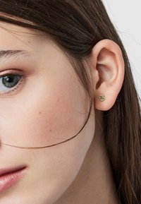 Pendiente de oro en forma de oso, con detalles de acento verde y una superficie pulida, llevado en el lóbulo de una oreja.