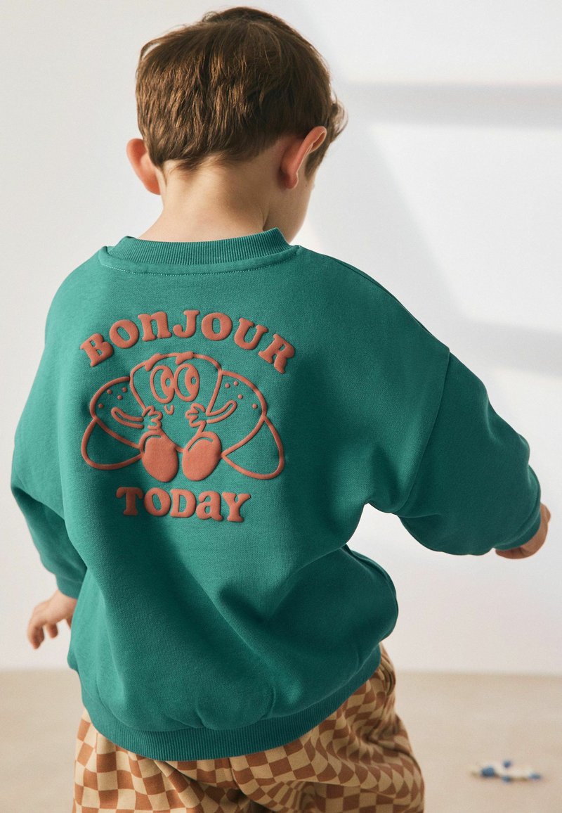 Felpa in teal con scollo rotondo, caratterizzata da una grafica in rilievo arancione con croissant in stile cartone animato e la scritta "BONJOUR TODAY" sul retro.