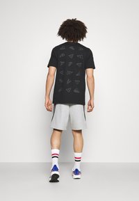 Camiseta negra con diseños de logotipos geométricos repetidos en la espalda, combinada con pantalones cortos gris claro y calcetines blancos con rayas rojas.