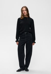 Jeune femme aux cheveux bruns détachés, portant un pull noir, un pantalon noir taille haute avec ceinture et des chaussures noires, debout devant un fond uni.