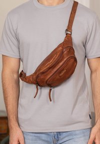 Borsa a tracolla in pelle marrone con due scomparti con zip, tracolla regolabile e forma curva, indossata su una maglietta grigia chiara.