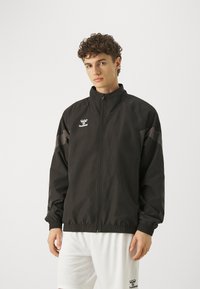 Svart idrottsjacka med dragkedja fram, hög krage och elastiska manschetter. Har logotyp och grå detaljer på ärmarna, tillverkad av lätt material.