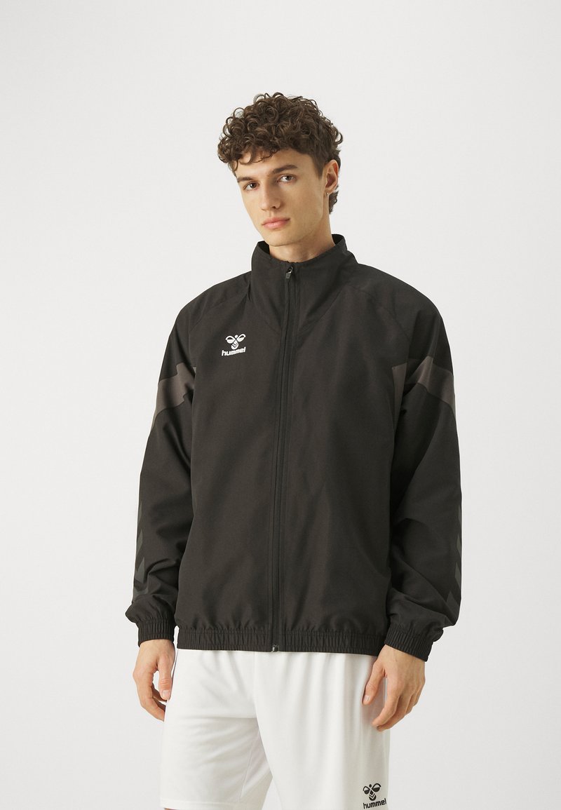 Svart idrottsjacka med dragkedja fram, hög krage och elastiska manschetter. Har logotyp och grå detaljer på ärmarna, tillverkad av lätt material.