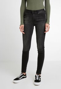 Pantalones vaqueros ajustados de mezclilla negra con un acabado ligeramente descolorido, acentuados por un diseño clásico de 5 bolsillos, combinados con zapatillas de lona negras y blancas.