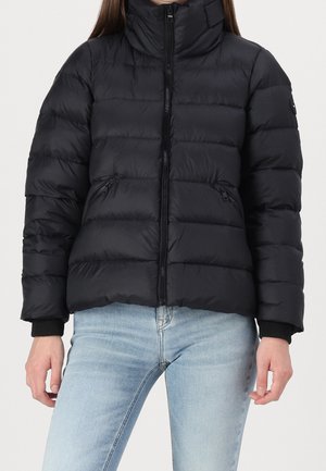 Vrouw draagt een zwart doorgestikt donzen jack met voorrits en lichtblauwe spijkerbroek tegen een effen achtergrond.