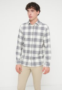 Chemise à carreaux blancs et bleu marine à boutons, manches longues, dotée d'un col et de boutons en bois, portée avec un pantalon beige clair.