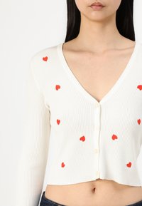 Cardigan bianco a coste con profondo scollo a V, sei bottoni e applicazioni a forma di cuore rosse sparse su tutto il tessuto. Design cropped.
