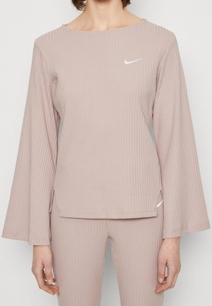 Kobieta w beżowym prążkowanym topie z długim rękawem i dopasowanych spodniach z białymi logo Nike Swoosh na piersi i biodrze.