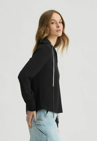 Zwarte top met lange mouwen en een V-hals, versierd met zilveren studs aan de voorkant; gecombineerd met lichtblauwe high-waisted jeans.
