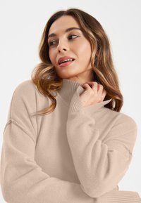 Beige trui met een hoge ribbelkraag, lange mouwen en een textuurstof. Het ontwerp heeft een losse pasvorm en subtiele stikseldetails.