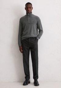 Grijze, ritssweater met hoge kraag, gecombineerd met donkere, gestructureerde broeken. Model draagt zwarte schoenen en staat tegen een neutrale achtergrond.