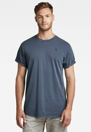 T-shirt in cotone blu con collo a giro e maniche arrotolate, caratterizzato da un piccolo logo sul petto e una vestibilità comoda. Pantaloni a righe visibili sotto.