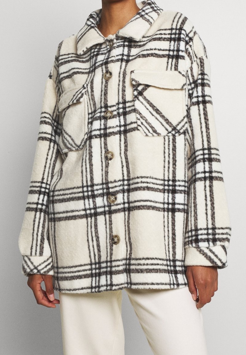 Personne portant une veste à carreaux blanche et noire à boutons avec deux poches poitrine et un pantalon blanc.