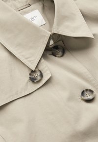 Beige trench coat collar with large, round, marbled buttons and visible stitching, showing part of the brand label inside.
