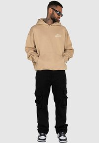 Beige hoodie met een voorzak en logo; gecombineerd met zwarte cargobroek met zijzakken; zwart-witte sneakers.